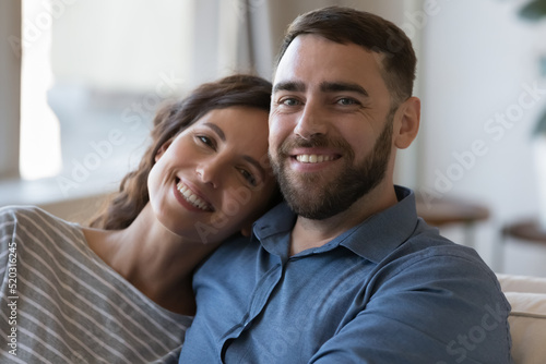Happy young adult married couple home head shot portrait. Husband and wife resting on couch, hugging in at home, looking at camera, smiling, laughing. Love, marriage, relationship concept