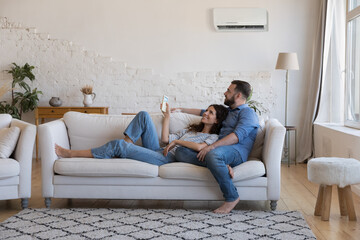 Happy millennial couple of homeowners enjoying cool conditioned air, resting on couch together, using remote control for AC, cooling domestic equipment start. Home appliance concept