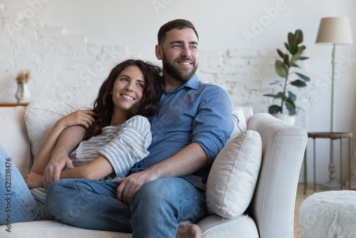 Cheerful dreamy young dating couple in love resting on sofa, relaxing at home, enjoying leisure, hugging, looking away, planning family future, marriage, mortgage, talking, laughing