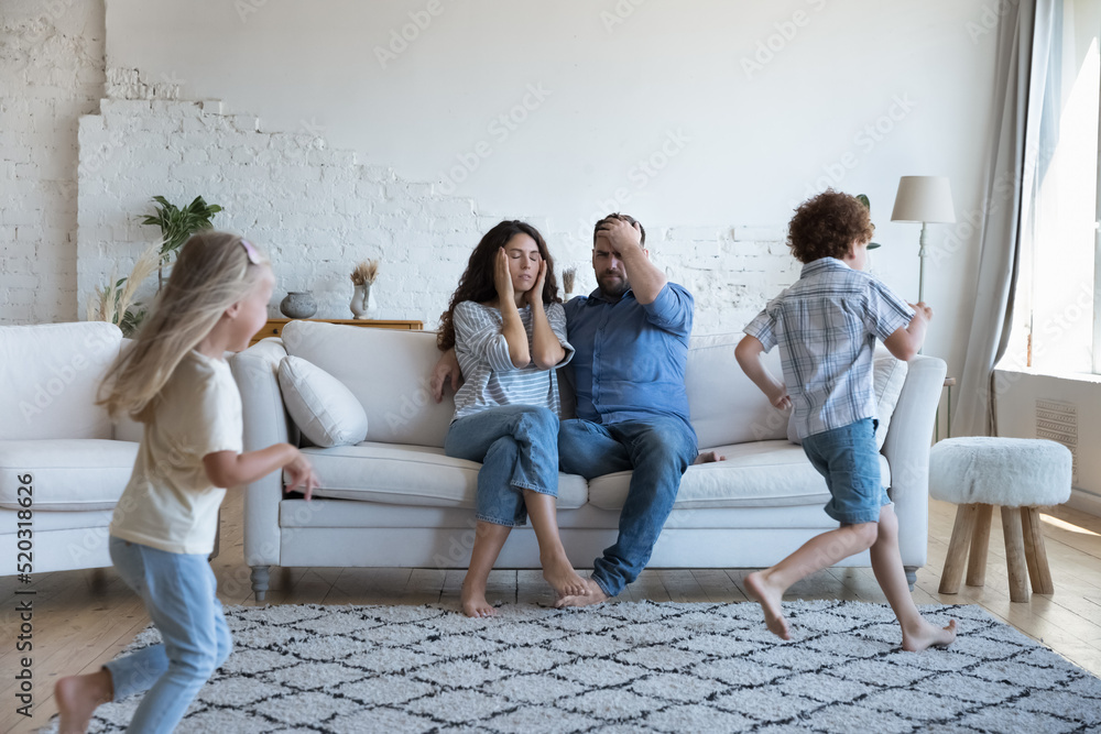 Fototapeta premium Two active sibling kids running around tired parents, playing active games. Mom and dad exhausted with watching children, feeling headache, stress. Upbringing problems, parenthood, family concept