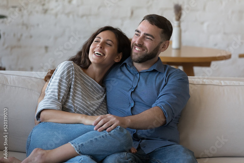 Happy young adult couple in love, enjoying leisure on couch, hugging, looking away, dreaming, thinking over marriage, family, real estate buying, talking, chatting, laughing