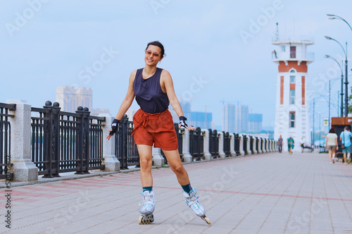 A young woman with rollerblades on the embankment in protective gloves and goggles. Sports lifestyle. The girl smiles, having fun. Lighthouse in blur in the background.