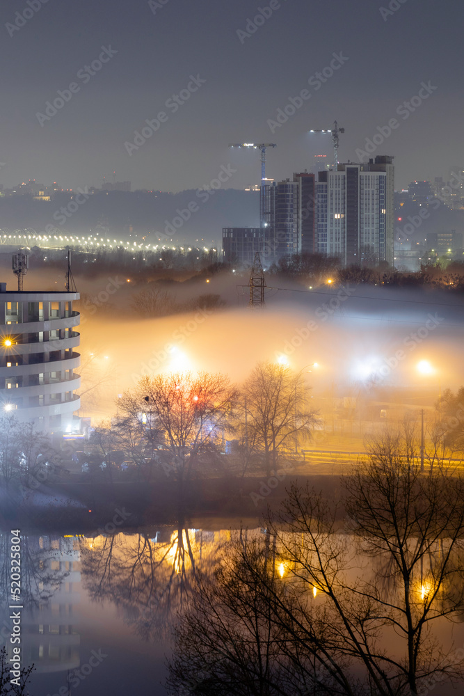 Night cityscape of the big city. Awesome bright, multi colored light in thick fog at empty streets. Apartment buildings in bedroom community, commuter town area. Darnytsia, Kyiv. Ukraine. 2021