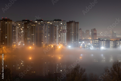 Night cityscape of the big city. Awesome bright, multi colored light in thick fog at empty streets. Apartment buildings in bedroom community, commuter town area. Darnytsia, Kyiv. Ukraine. 2021 photo