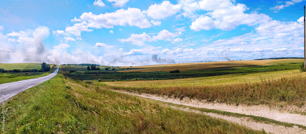 Countryside panoramic view with asphalt road, fields, groves, hills, sky with clouds. Ukraine summer landscape in Lviv Region. Smoke over the field of wheat due to fire in background. Scenic landscape