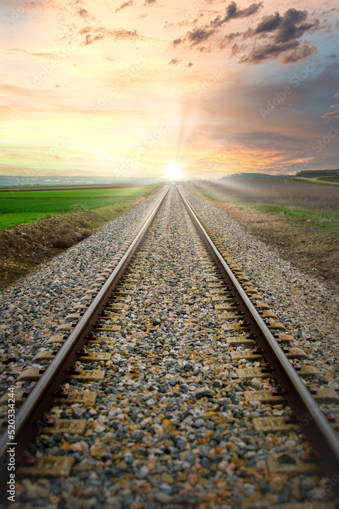 Vías del tren a través de campos de cultivo, que convergen hacia el sol en el horizonte - obrazy, fototapety, plakaty 
