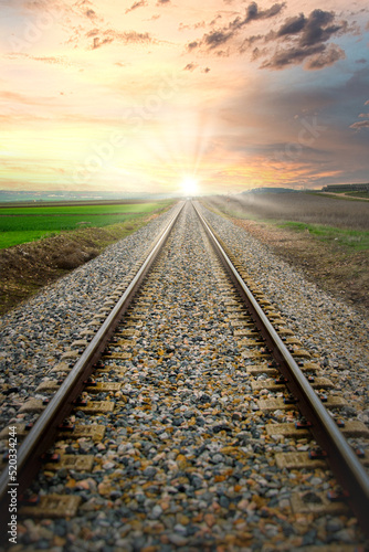 Vías del tren a través de campos de cultivo, que convergen hacia el sol en el horizonte