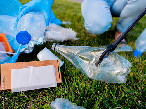 Volunteers clean up trash from the lawn in the alley. Tongs for collecting garbage hold a dirty plastic bottle. Household waste garbage bag