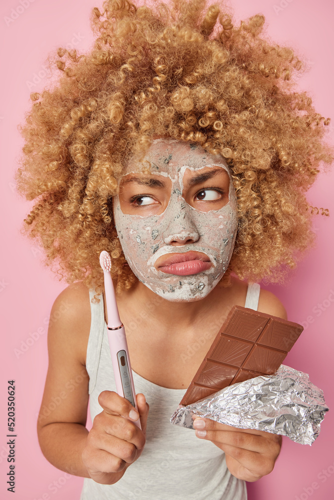 Unhappy woman with curly hair holds electric toothbrush and bar of chocolate applies beauty nourishing mask on face for skin treatment isolated over pink background. Beauty and oral hygiene. Unhappy woman with curly hair holds electric toothbrush and bar of chocolate applies beauty nourishing mask on face for skin treatment isolated over pink background. Beauty and oral hygiene.