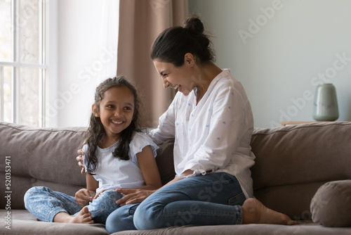 Cheerful Indian mom cuddling happy little daughter on couch at home with love, care, gratitude, talking to kid, smiling, laughing, having fun, enjoying motherhood, family leisure time
