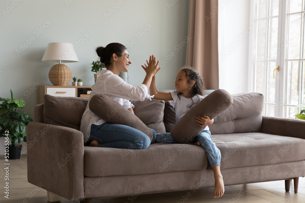 Happy mom and cheerful little kid giving high five after pillow fighting battle, sitting on couch, holding cushions, clapping hands, laughing, talking, playing active games. Family leisure concept Happy mom and cheerful little kid giving high five after pillow fighting battle, sitting on couch, holding cushions, clapping hands, laughing, talking, playing active games. Family leisure concept