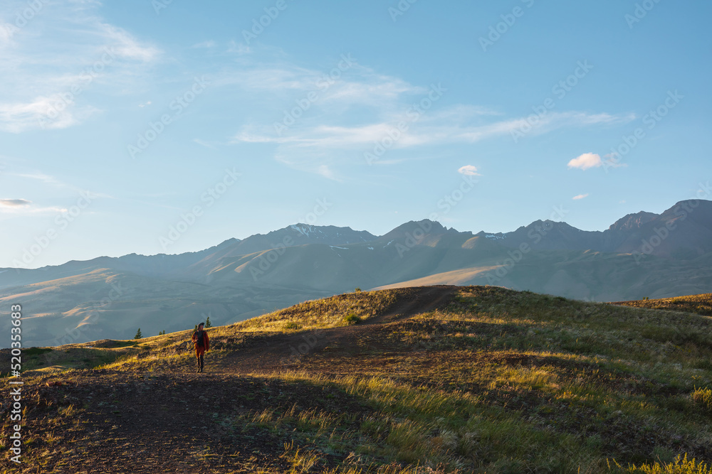 Tourist on sunlit gold grassy hill with dirt road with view to high mountain range. Beautiful sunny mountain landscape with man in mountains. Colorful scenery with traveler on hill in golden sunlight.