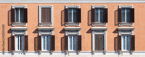 Photography Shuttered windows, some closed, in an apartment building with an orange plastered wall on a sunny day in Rome, Italy