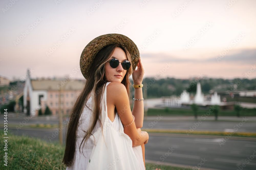 Portrait of a relaxed happy woman in white dress with hat looking at the camera. Cityscape in the background