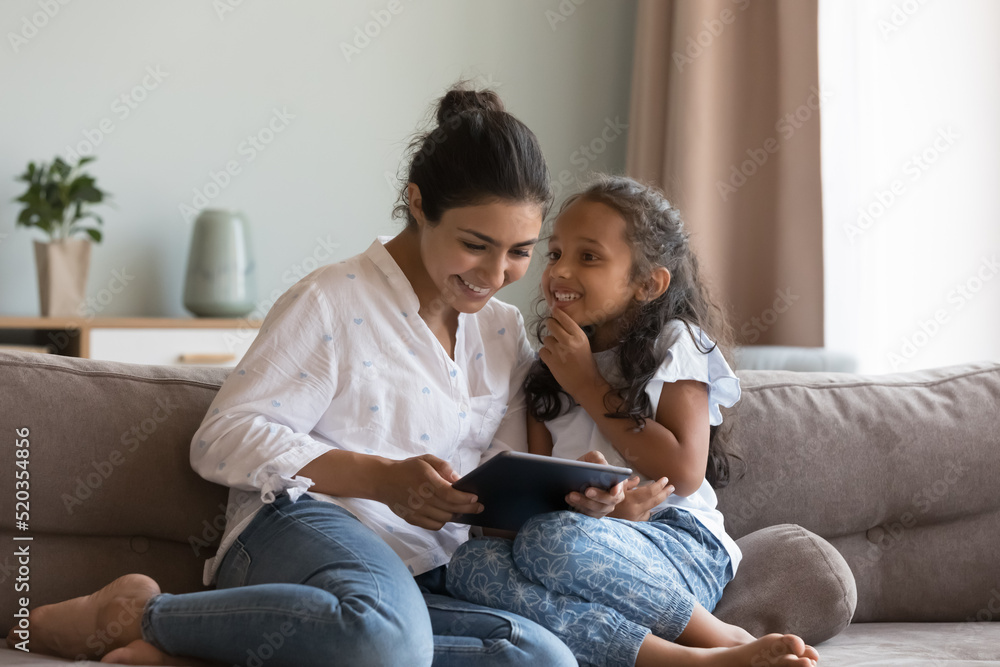Happy Indian little daughter kid talking to mom, using digital tablet, sitting on sofa at home, smiling, laughing. Mother and child enjoying leisure, playing game on digital gadget Happy Indian little daughter kid talking to mom, using digital tablet, sitting on sofa at home, smiling, laughing. Mother and child enjoying leisure, playing game on digital gadget