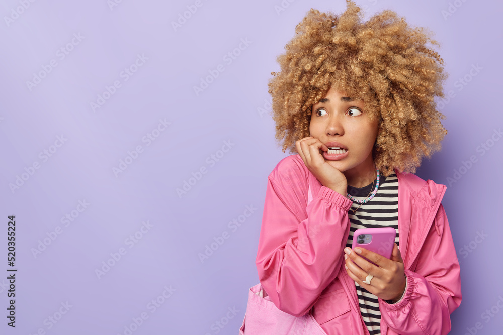 Worried concerned young woman bites finger nails looks anxious aside holds mobile phone has problem receiving notification on cellular about bug dressed casually isolated over purple background
