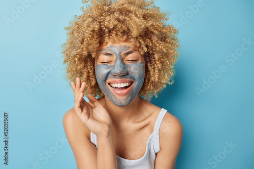 Horizontal shot of positive curly haired woman applies facial mud mask to inscrease hydration and revitalize demaged skin enjoys pampering procedures smiles broadly wears t shirt poses indoors photo