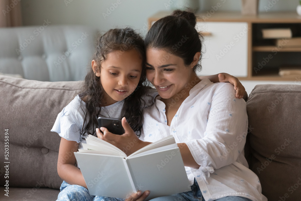 Happy Indian mom hugging little kid on couch, showing smartphone screen to girl on couch, using online app, browsing Internet. Mum and daughter reading paper book at home Happy Indian mom hugging little kid on couch, showing smartphone screen to girl on couch, using online app, browsing Internet. Mum and daughter reading paper book at home