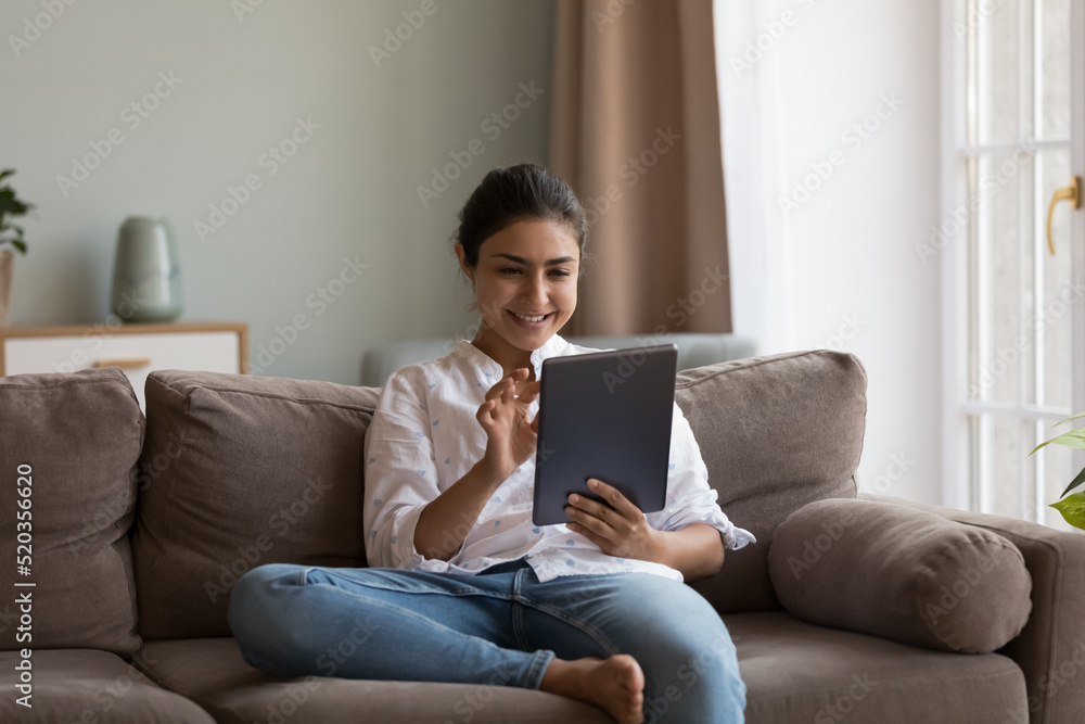 Happy millennial Indian girl using online app on digital device at home, reading book, ebook on tablet, browsing internet, social media, shopping on webstores, resting on soft couch at home Happy millennial Indian girl using online app on digital device at home, reading book, ebook on tablet, browsing internet, social media, shopping on webstores, resting on soft couch at home