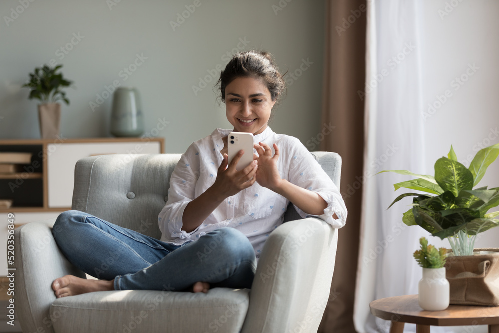 Happy pretty millennial Indian girl relaxing at home, resting in armchair, typing on smartphone, using online app, software, shopping on Internet, making video call. Mobile phone communication concept Happy pretty millennial Indian girl relaxing at home, resting in armchair, typing on smartphone, using online app, software, shopping on Internet, making video call. Mobile phone communication concept