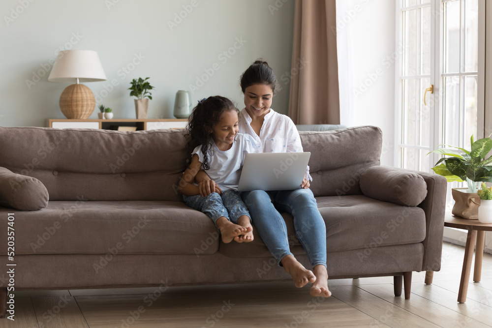 Happy pretty Indian mother showing learning app on laptop to little daughter. Mom and cute girl watching movie together, talking on video call, enjoying leisure in home living room cozy interior Happy pretty Indian mother showing learning app on laptop to little daughter. Mom and cute girl watching movie together, talking on video call, enjoying leisure in home living room cozy interior