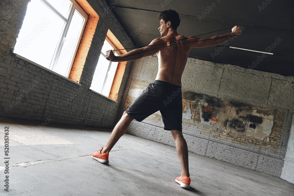 Full length of concentrated young man exercising with resistance band in gym Full length of concentrated young man exercising with resistance band in gym