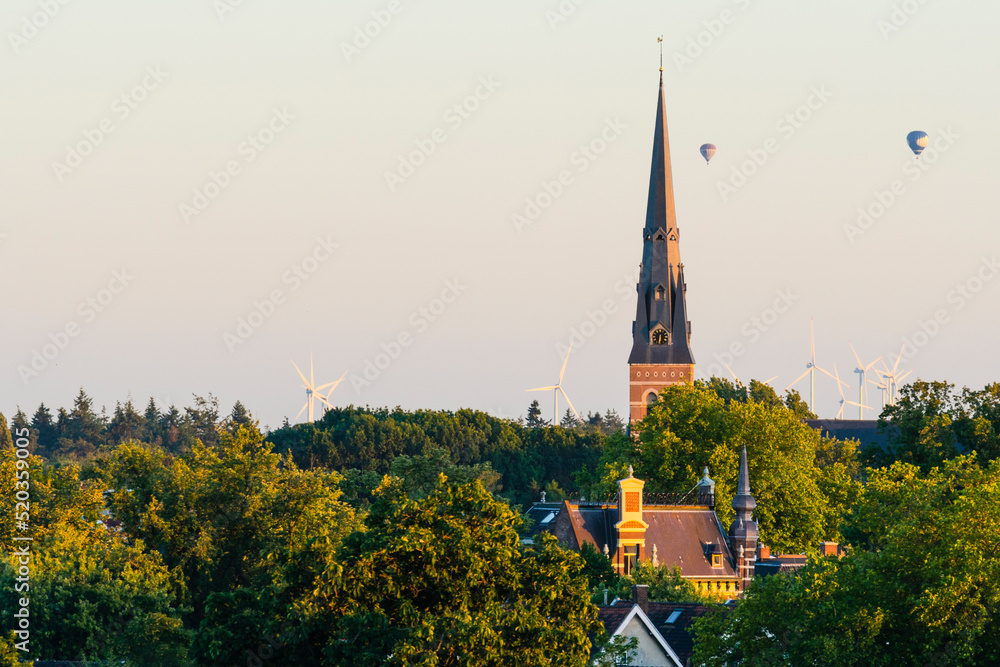 European cathedral tower surrounded by green trees at sunset with hot air balloons. Breda, the netherlands European cathedral tower surrounded by green trees at sunset with hot air balloons. Breda, the netherlands