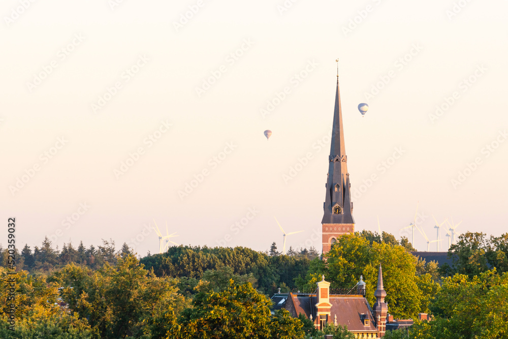 Cityscape small northern european city. European cathedral tower surrounded by green trees at sunset with hot air balloons. Breda, the netherlands