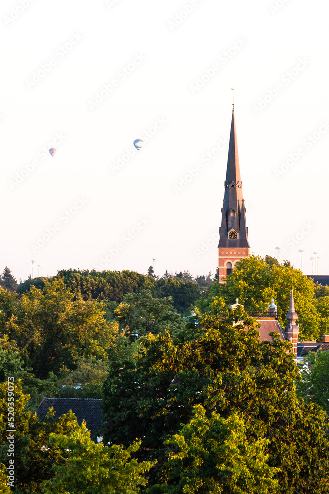 European cathedral tower surrounded by green trees at sunset with hot air balloons. Breda, the Netherlands