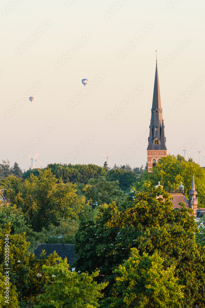 European cathedral tower surrounded by green trees at sunset with hot air balloons. Breda, the Netherlands