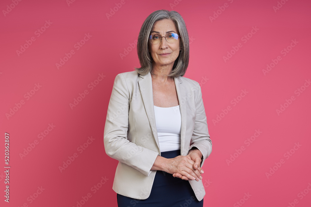 Confident senior woman in formalwear looking at camera while standing against pink background Confident senior woman in formalwear looking at camera while standing against pink background