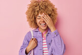 Cheerful curly haired woman makes face palm giggles happily dressed in purple jacket expresses positive emotions cannot stop laughing at joke isolated over pink background. Sincere feelings concept