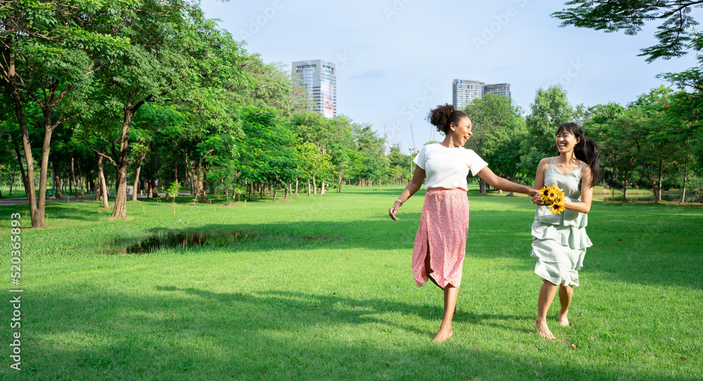 Friend girl concept.Two smiling girls run at green grass at background of blue sky with clouds.Young adult African Asian American woman walk on summer field. Empty space for inscription or objects.