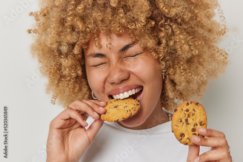 Εκτύπωση καμβά Close up shot of blonde curly haired woman eats cookies with chocolate bites deicious snack keeps eyes closed has sweet tooth dressed casually isolated over white background