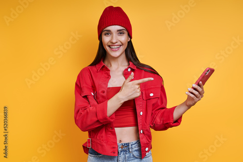 Happy young woman holding smart phone and covering mouth with hand against yellow background