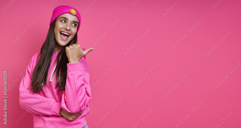 Beautiful woman in funky hat pointing away while standing against pink background Beautiful woman in funky hat pointing away while standing against pink background
