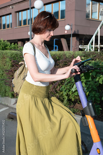 Middle-aged woman with a backpack rents an electronic scooter through a mobile application. City electric scooter rental service - kick sharing. Travel around the city on a scooter photo