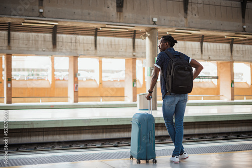 African male standing next to a platform while holding a blue suitcase. He is at a train station