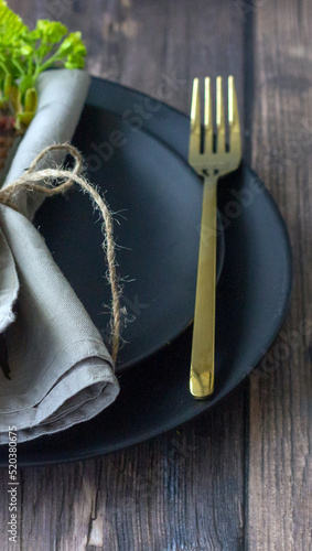 Table setting with black ceramic plate, golden fork, linen napkin on the wooden table.