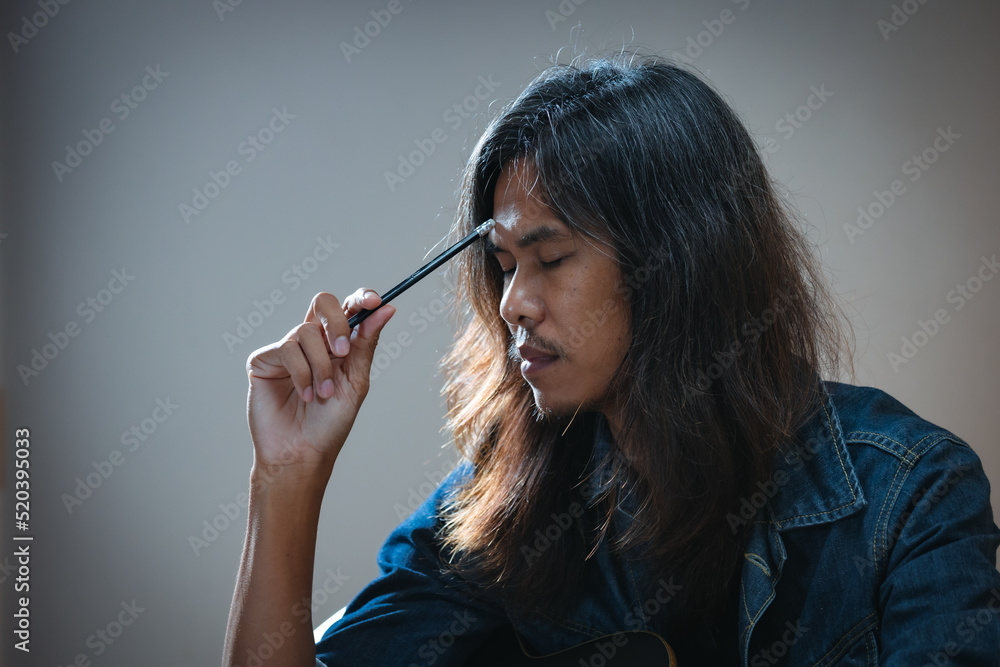 Obraz premium Asian musician who have long hair and wear jean jacket is playing guitar and so stressed to think about lyric and write a song in notebooks in the dark room.
