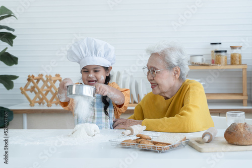 Happy family Asian elderly grandmother and little cute grandchild spend time together at kitchen, sift flour on dough for bake cookies or bread, smile and have fun, flour mess up on nose and face