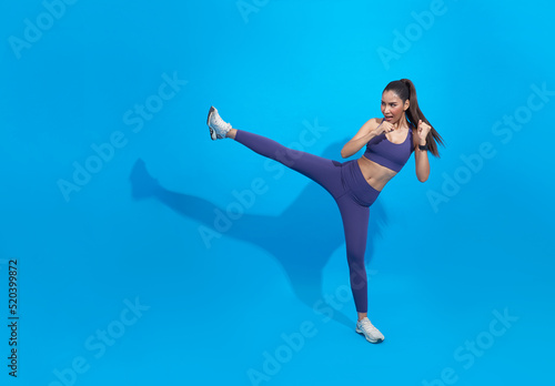 Asian female athlete workout, practice leg kicks, kicking air in sportswear. Muscular trained woman kicking with raised feet, exercise kickboxing moves, blue background.