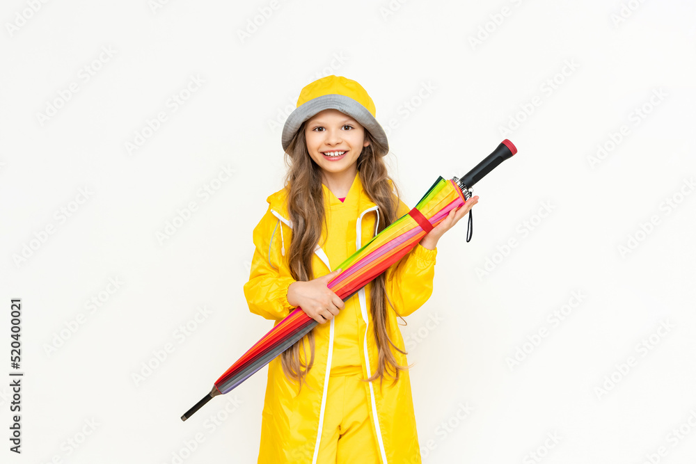 A beautiful girl holds a multicolored closed umbrella on a white isolated background. A child dressed in a yellow raincoat and a panama hat from the rain. Rainy weather concept. A beautiful girl holds a multicolored closed umbrella on a white isolated background. A child dressed in a yellow raincoat and a panama hat from the rain. Rainy weather concept.