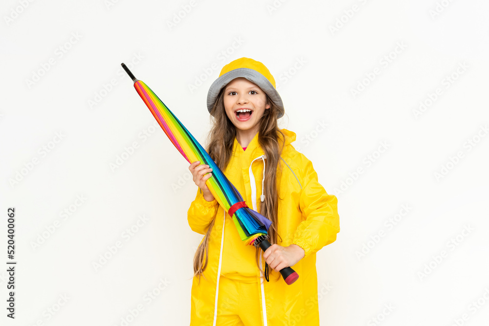 A beautiful girl holds a multicolored closed umbrella on a white isolated background. A child dressed in a yellow raincoat and a panama hat from the rain. A beautiful girl holds a multicolored closed umbrella on a white isolated background. A child dressed in a yellow raincoat and a panama hat from the rain.