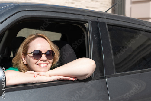 Smiling girl in sunglasses looking out the window sitting in a car in the parking lot, illuminated by the sun. The concept of traveling by car. Urban tourism