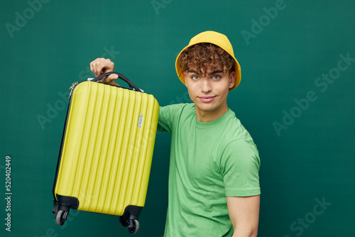 a cute, happy man stands on a green background, dressed in a yellow panama hat, holding a bright travel suitcase in his hand, lifting it.