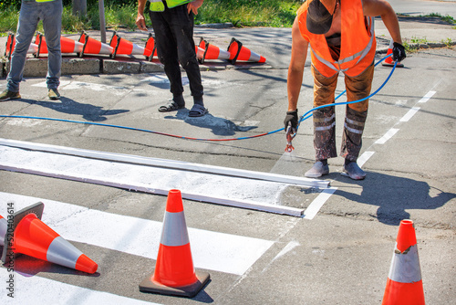 Slika na platnu A team of road workers paints a white road marking of a pedestrian crossing using a wooden frame template and an airbrush