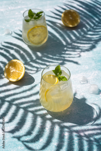 Homemade lemonade with lemon, mint and ice cubes in glasses on a light blue background. Cold summer refreshing drinks. Photo of lemonade with hard shadows.