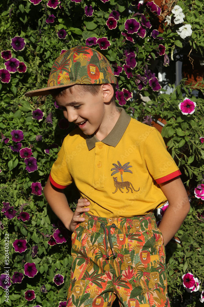 Fashion kid, boy, child in Moscow city, Russia. Summer stylish look for child, fashionable kid. Street style. Surfinia, petunia flowers in floral garden. Cute child