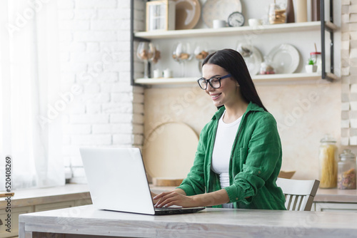 Young beautiful business woman in glasses and green shirt working at home in kitchen on laptop, smiling brunette working online remotely